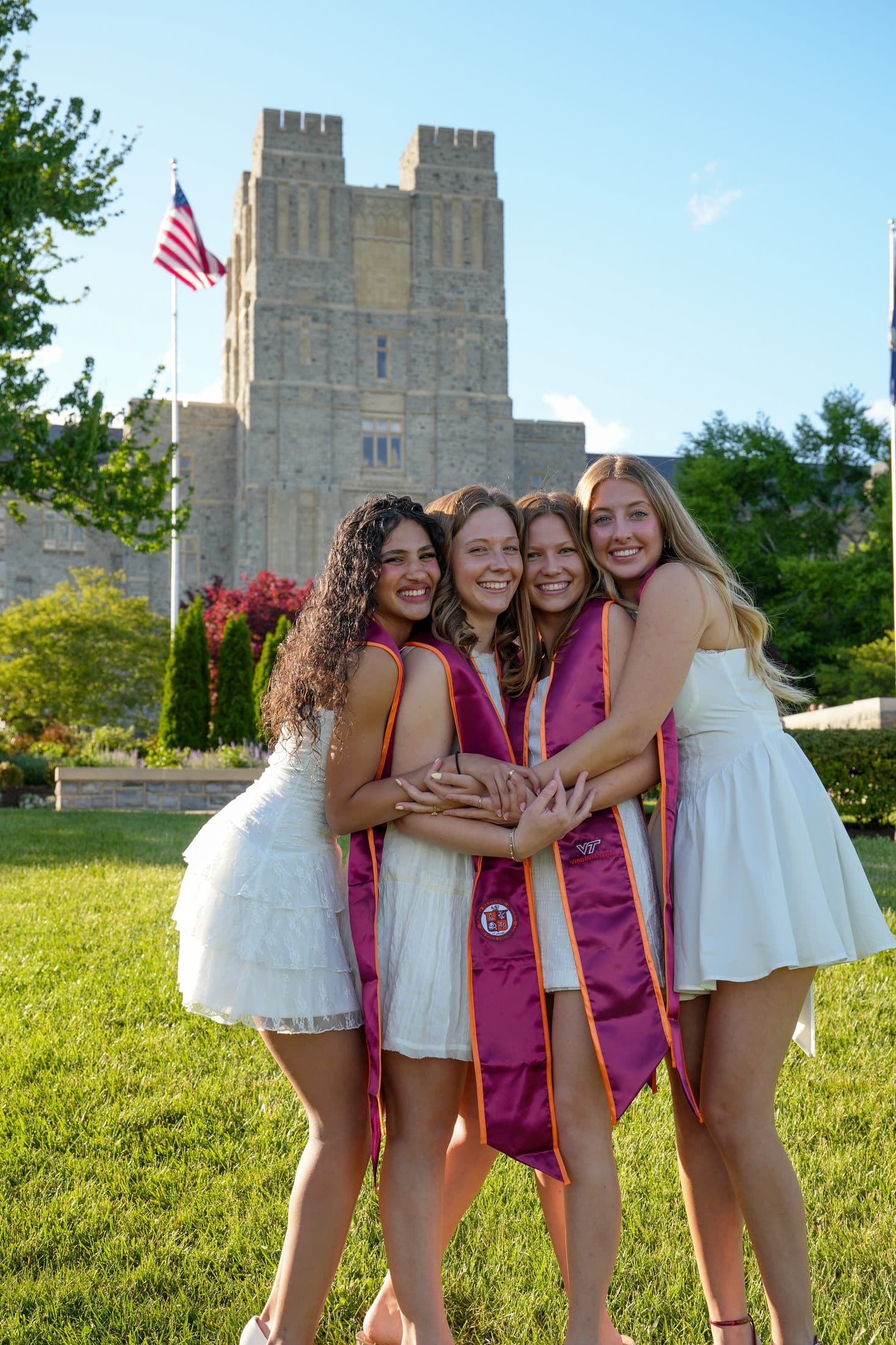 Four friends in white dresses and maroon stoles embracing below the campus tower