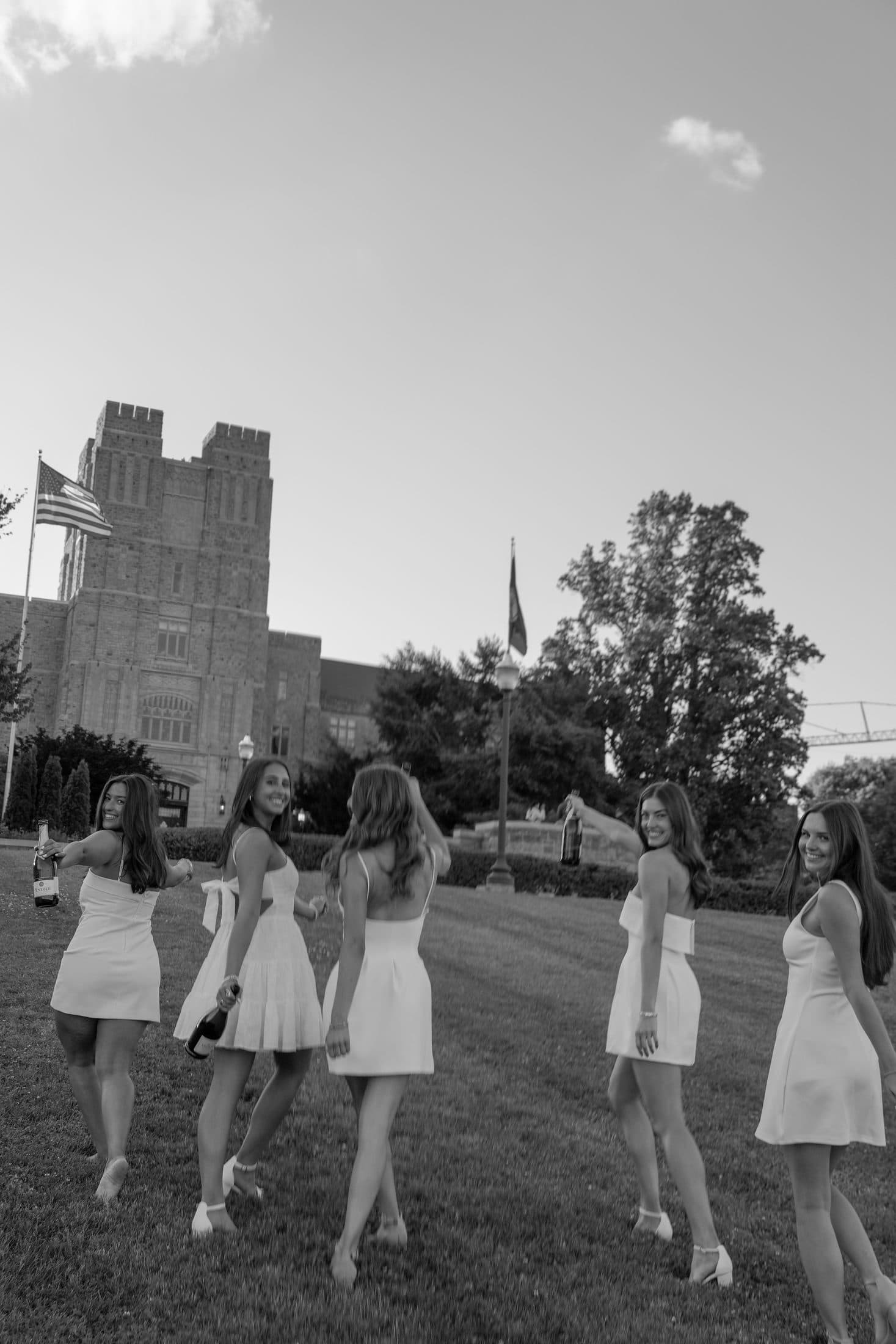 A line of graduates in white dresses walking past the campus flag