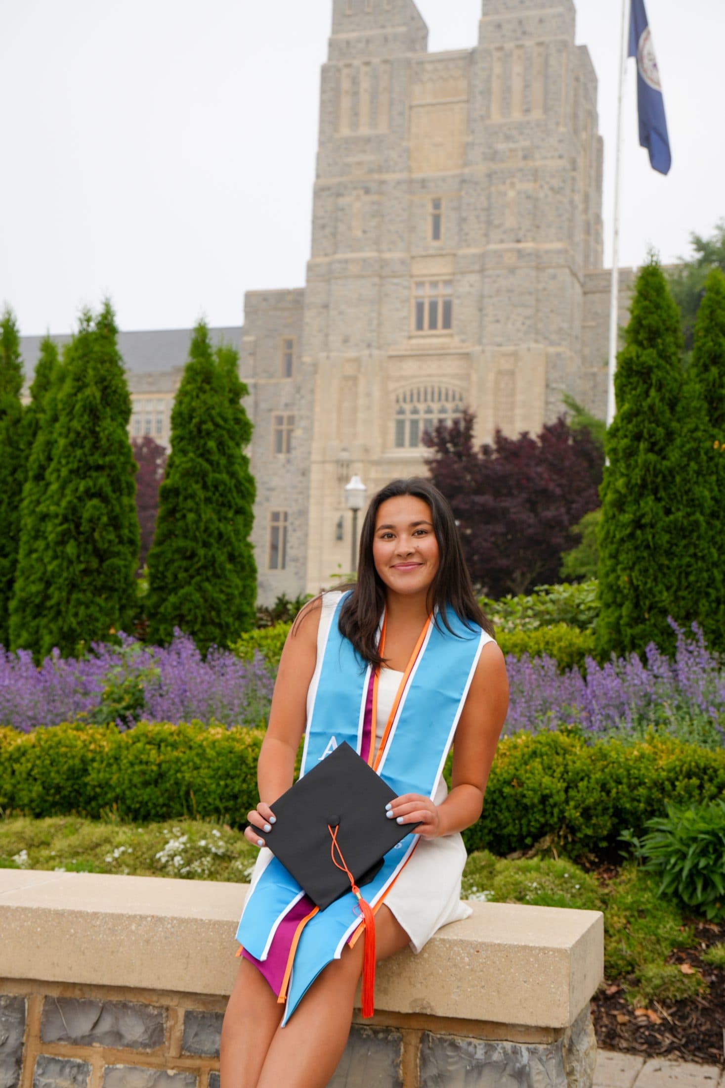 A graduate in a blue stole perched on a stone wall with the campus tower behind her