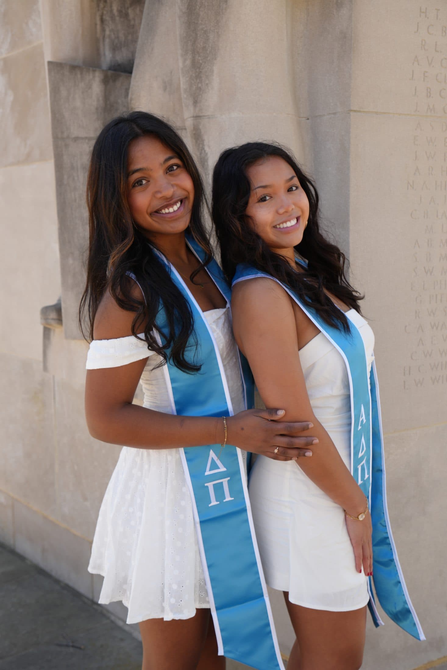 Two seniors in blue sorority stoles leaning against a stone pillar