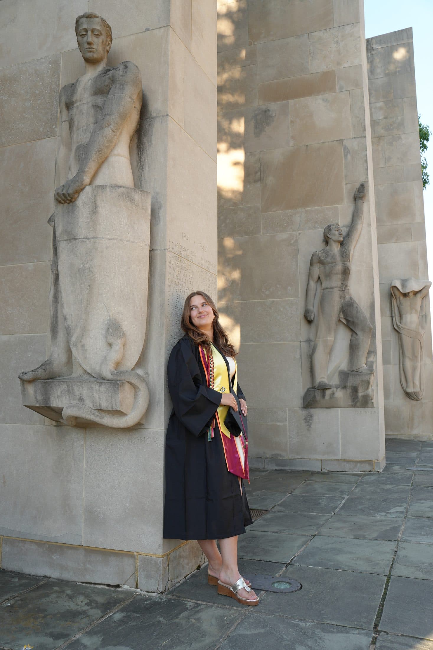A graduate in a black gown gazing up at the towering War Memorial statues