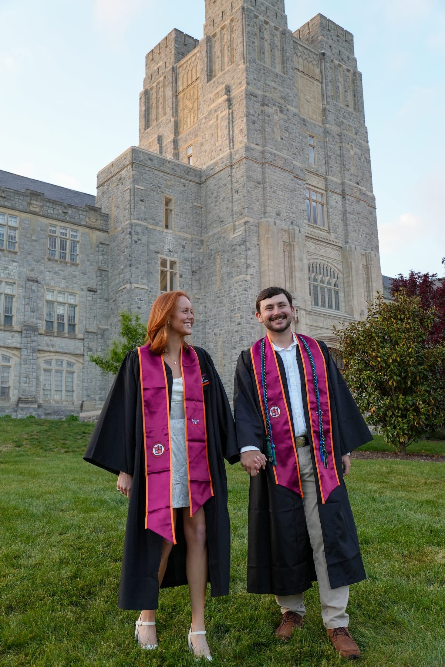 A couple in their gowns holding hands before Burruss Hall at dusk